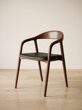 A mid-century modern wooden dining chair with a curved backrest, armrests, and a black upholstered seat, shown on a light wood floor against a neutral beige wall.