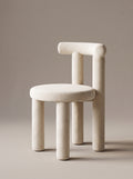 A minimalist, sculptural stool with a rounded, cushioned seat and thick cylindrical legs, upholstered in a warm off-white fabric, set against a solid taupe background.