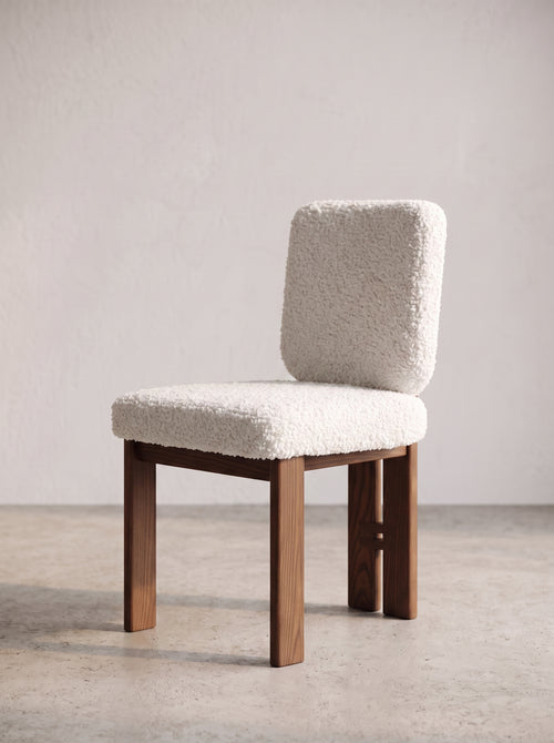 A modern side chair with a textured cream bouclé upholstered seat and backrest, supported by a medium brown wooden frame, set against a neutral concrete floor and textured plaster wall.