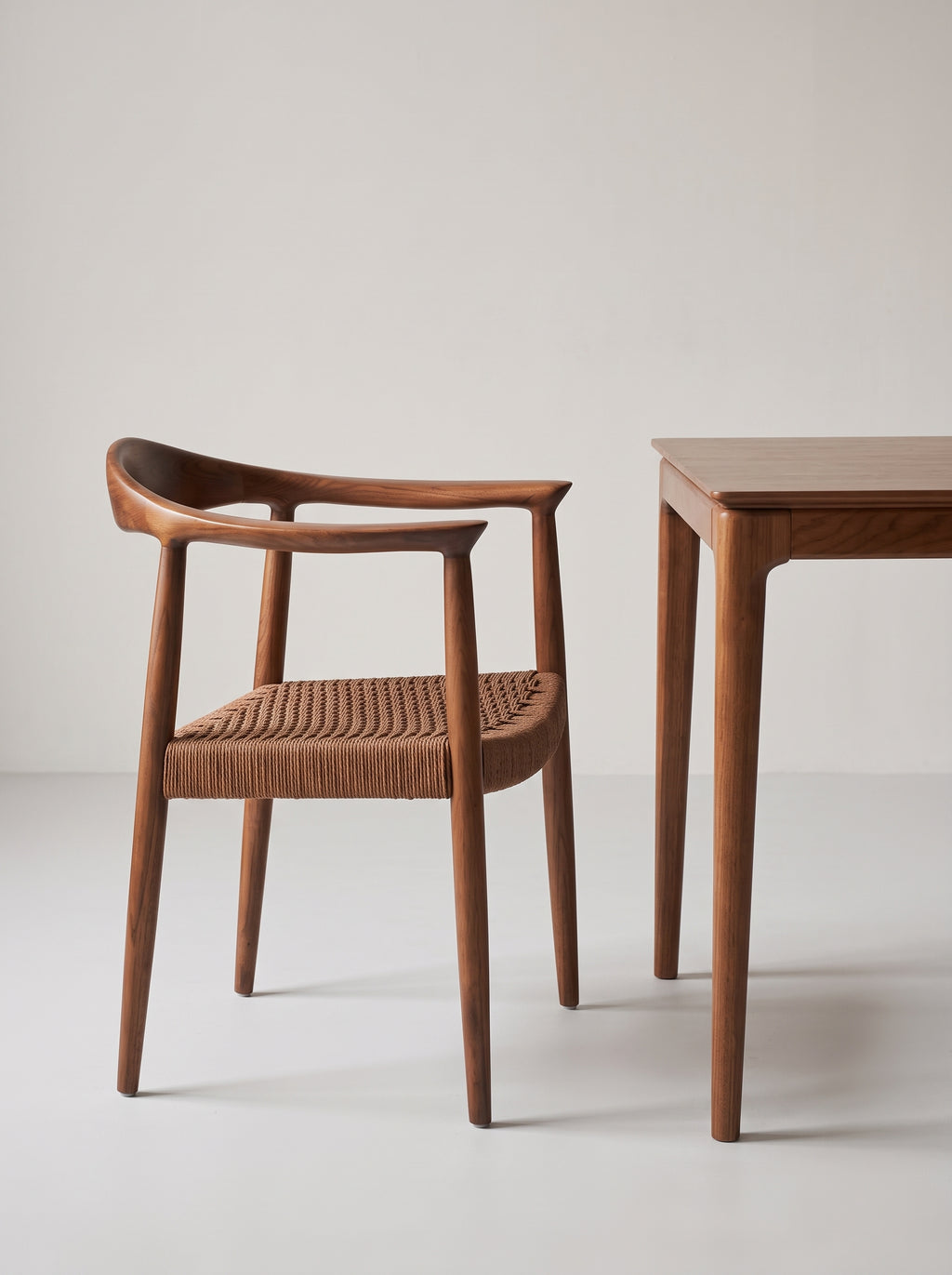 A mid-century modern wooden dining chair with a curved armrest and woven cane seat, shown next to a matching wooden table against a neutral light gray background.