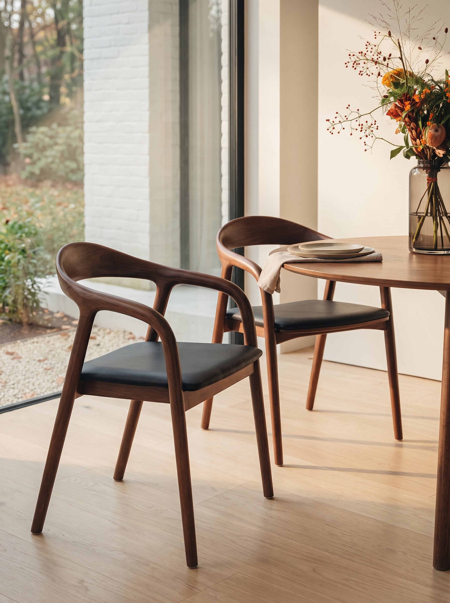 Two mid-century modern wooden dining chairs with black upholstered seats, shown in a bright dining room setting next to a wooden table with a vase of flowers.