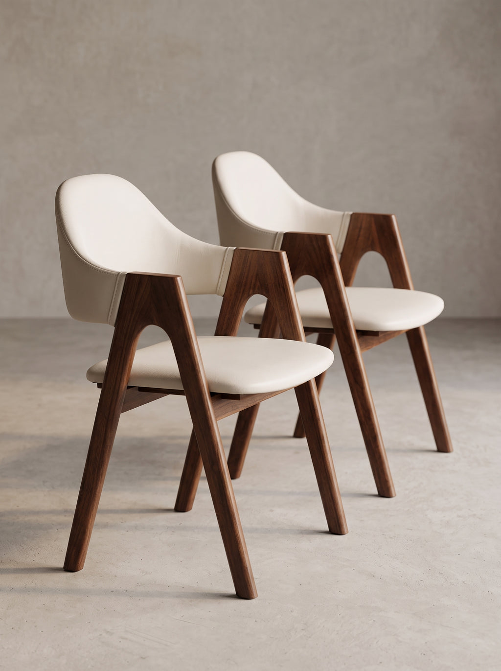 Two modern dining chairs with cream-colored upholstered seats and curved wooden legs, arranged on a concrete floor against a textured gray wall.