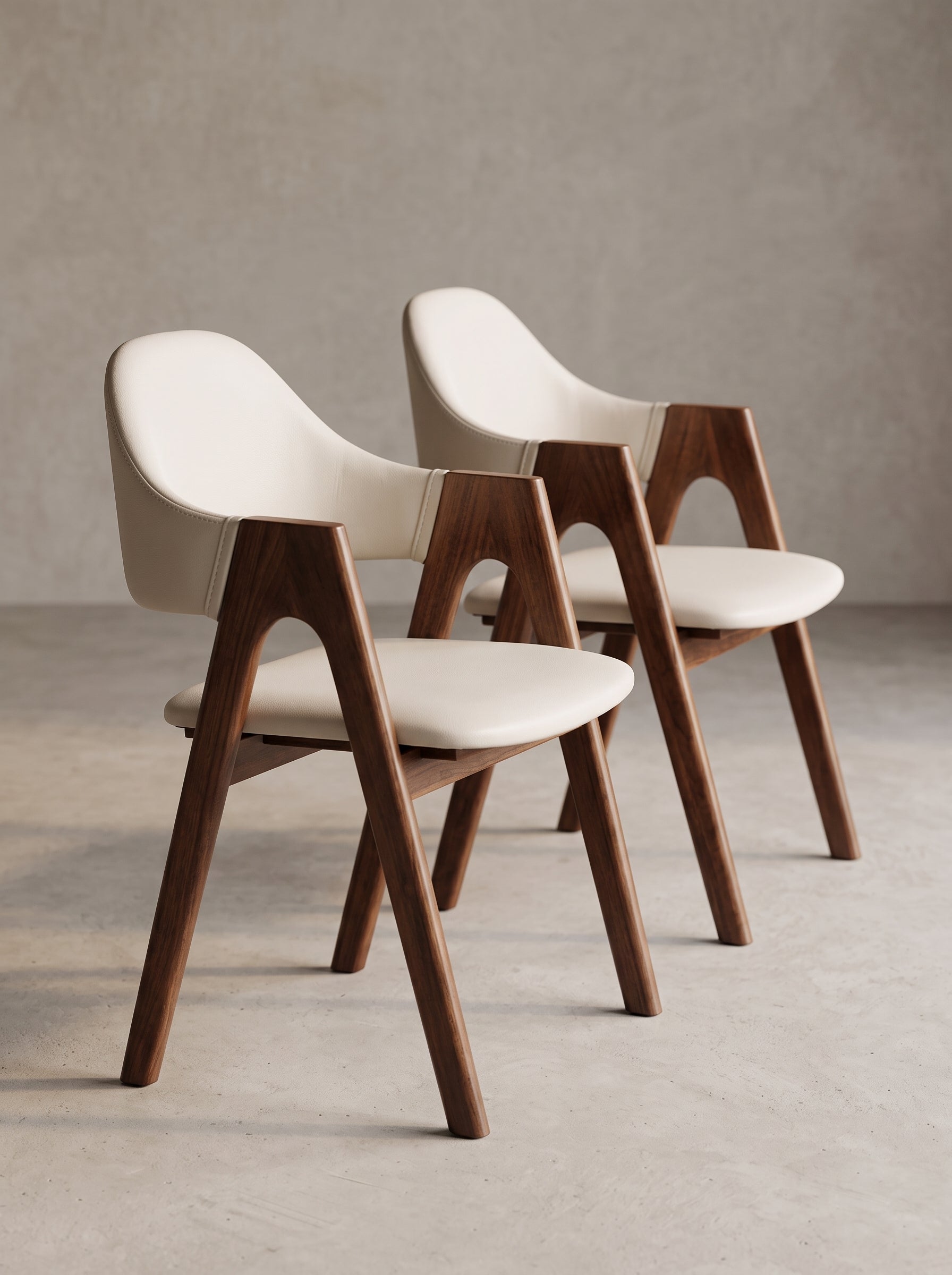 Two modern dining chairs with cream-colored upholstered seats and curved wooden legs, arranged on a concrete floor against a textured gray wall.