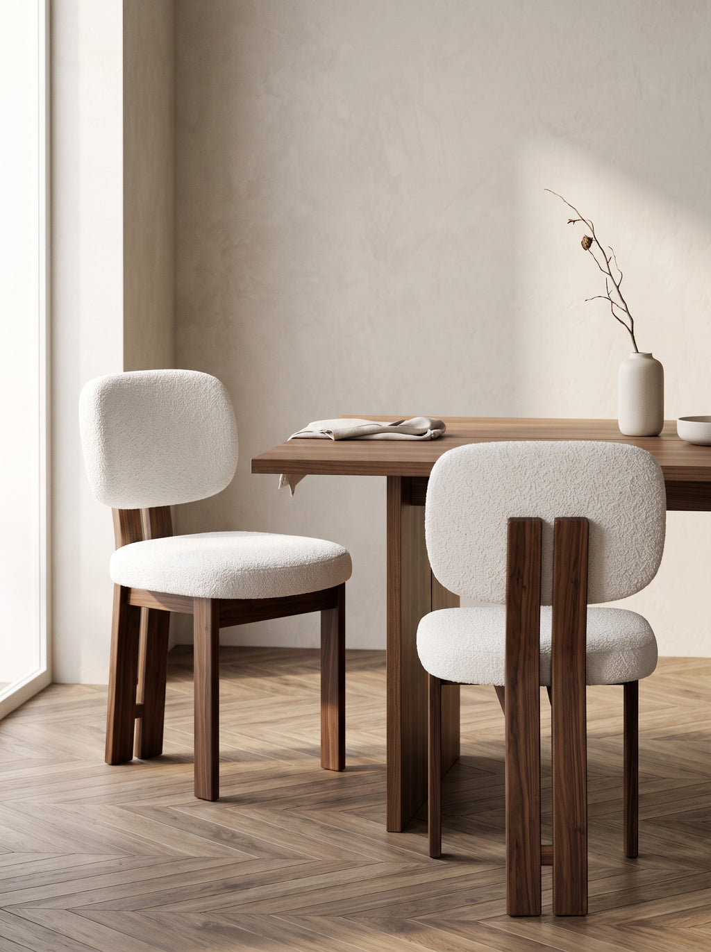 Two modern dining chairs with textured off-white bouclé upholstery and dark walnut wood legs, positioned around a matching wooden dining table in a minimalist room with a textured beige wall and herringbone wood flooring.