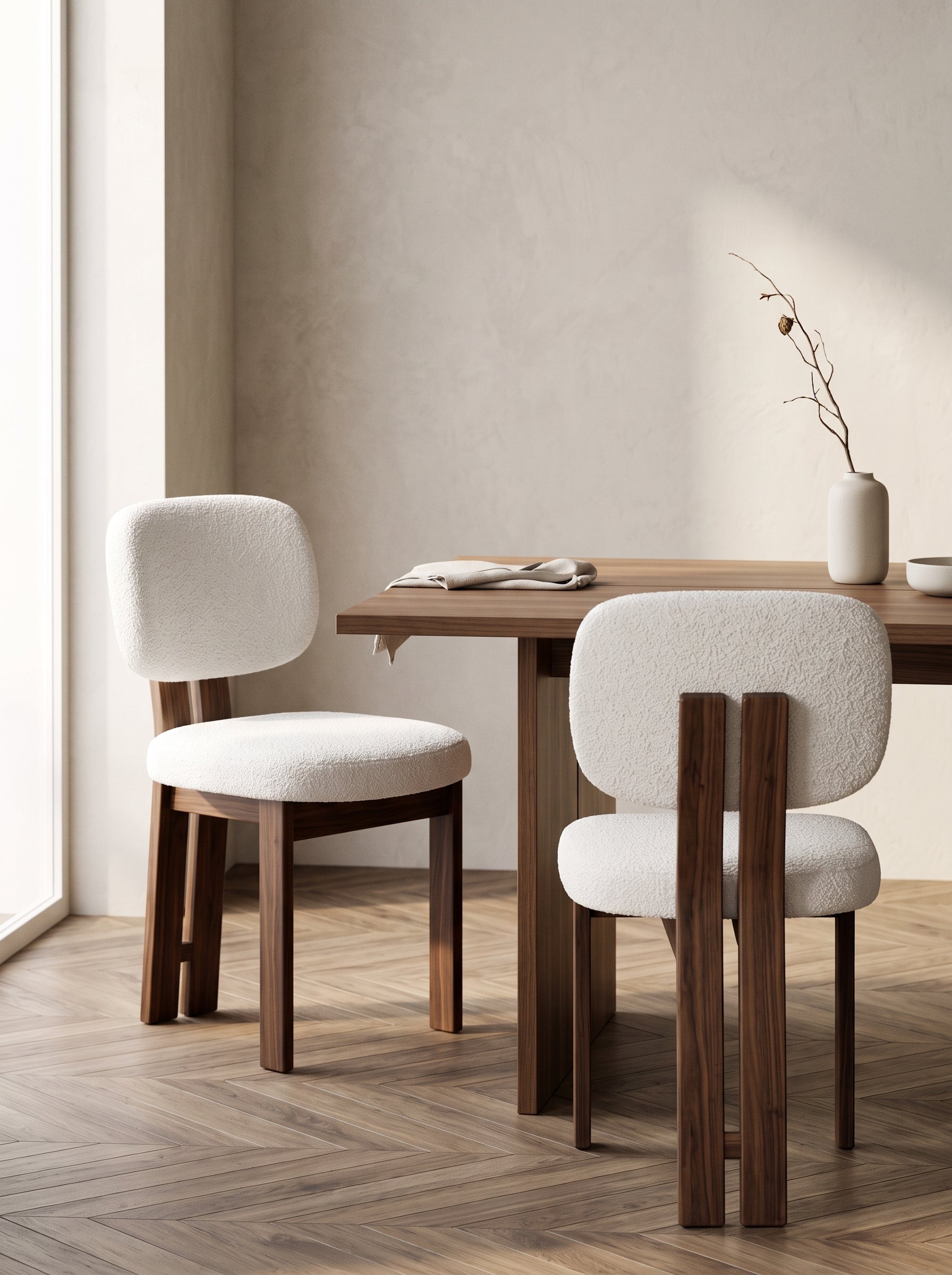 Two modern dining chairs with textured off-white bouclé upholstery and dark walnut wood legs, positioned around a matching wooden dining table in a minimalist room with a textured beige wall and herringbone wood flooring.