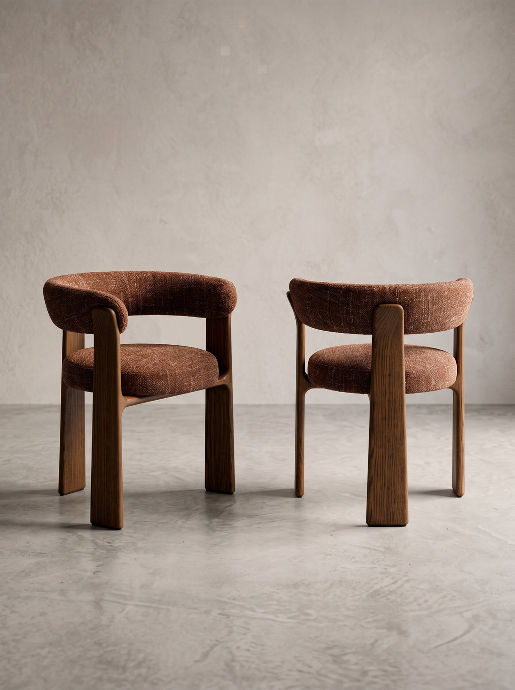 Two modern upholstered dining chairs with curved brown wood frames and textured brown fabric seats, set against a neutral concrete wall and floor.