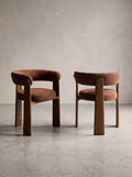 Two modern upholstered dining chairs with curved brown wood frames and textured brown fabric seats, set against a neutral concrete wall and floor.