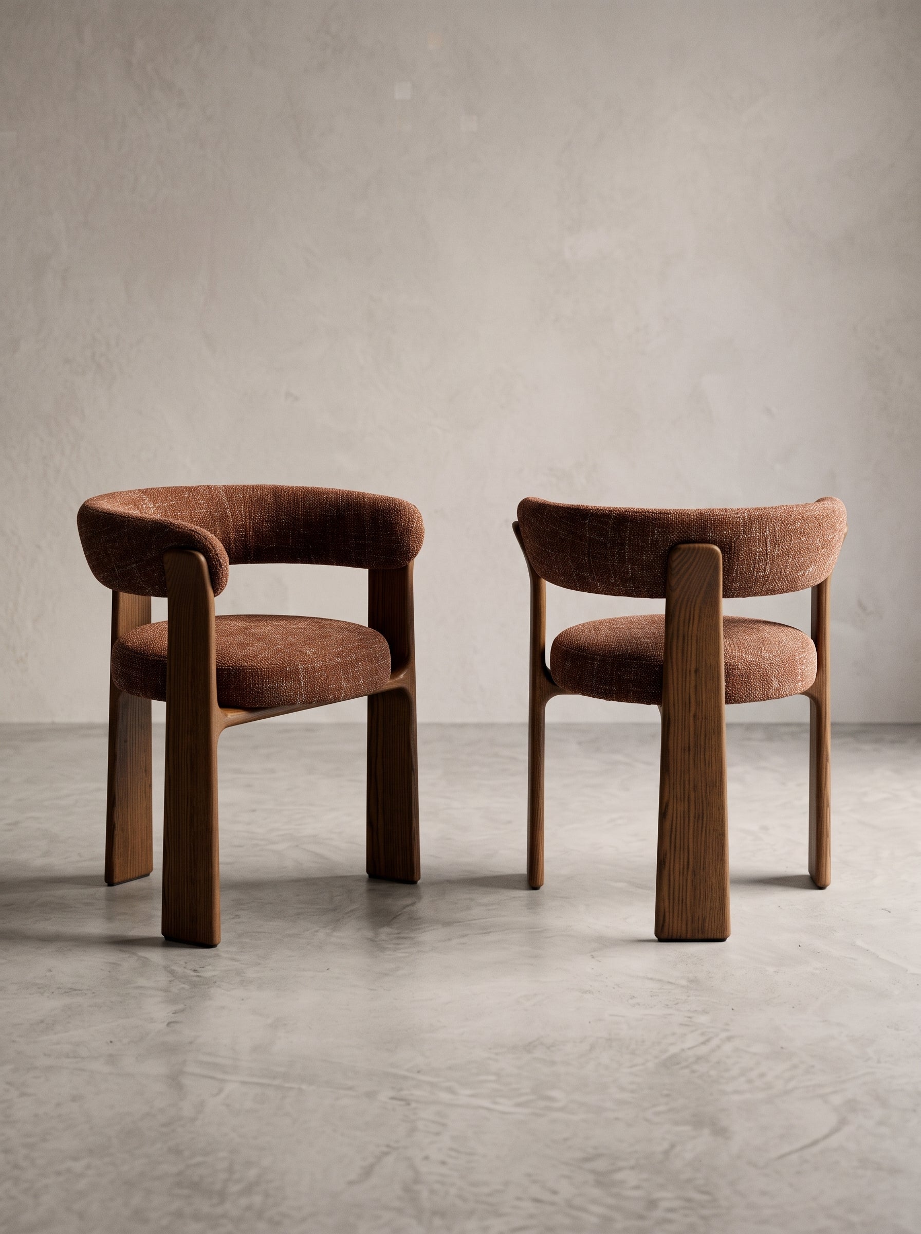 Two modern upholstered dining chairs with curved brown wood frames and textured brown fabric seats, set against a neutral concrete wall and floor.