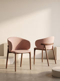 Two modern upholstered dining chairs with dusty pink curved seats and light brown wooden legs, set against a minimalist beige wall with soft diagonal shadows.