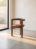 A modern wooden dining chair with a curved backrest and a brown leather seat cushion, positioned in a minimalist room with a concrete floor, textured beige plaster wall, and a large window on the left.