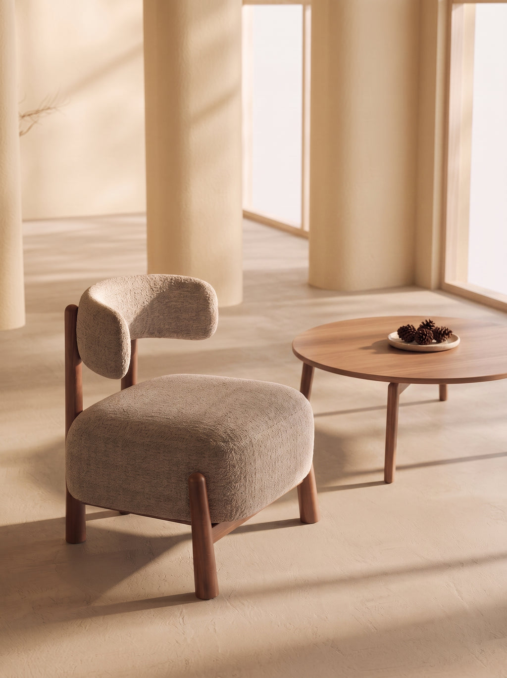 A modern upholstered accent chair with a curved backrest and wooden legs, shown beside a round wooden coffee table in a warm, sunlit interior with cream columns.