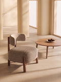 A modern upholstered accent chair with a curved backrest and wooden legs, shown beside a round wooden coffee table in a warm, sunlit interior with cream columns.
