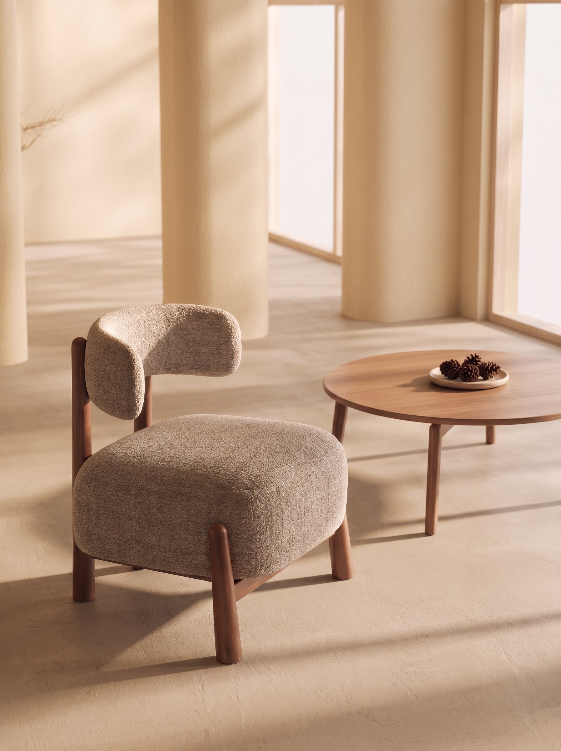 A modern upholstered accent chair with a curved backrest and wooden legs, shown beside a round wooden coffee table in a warm, sunlit interior with cream columns.
