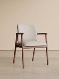 A mid-century modern armchair with a light beige upholstered seat and backrest, supported by a warm brown wooden frame, set against a minimalist beige wall and light wood floor.