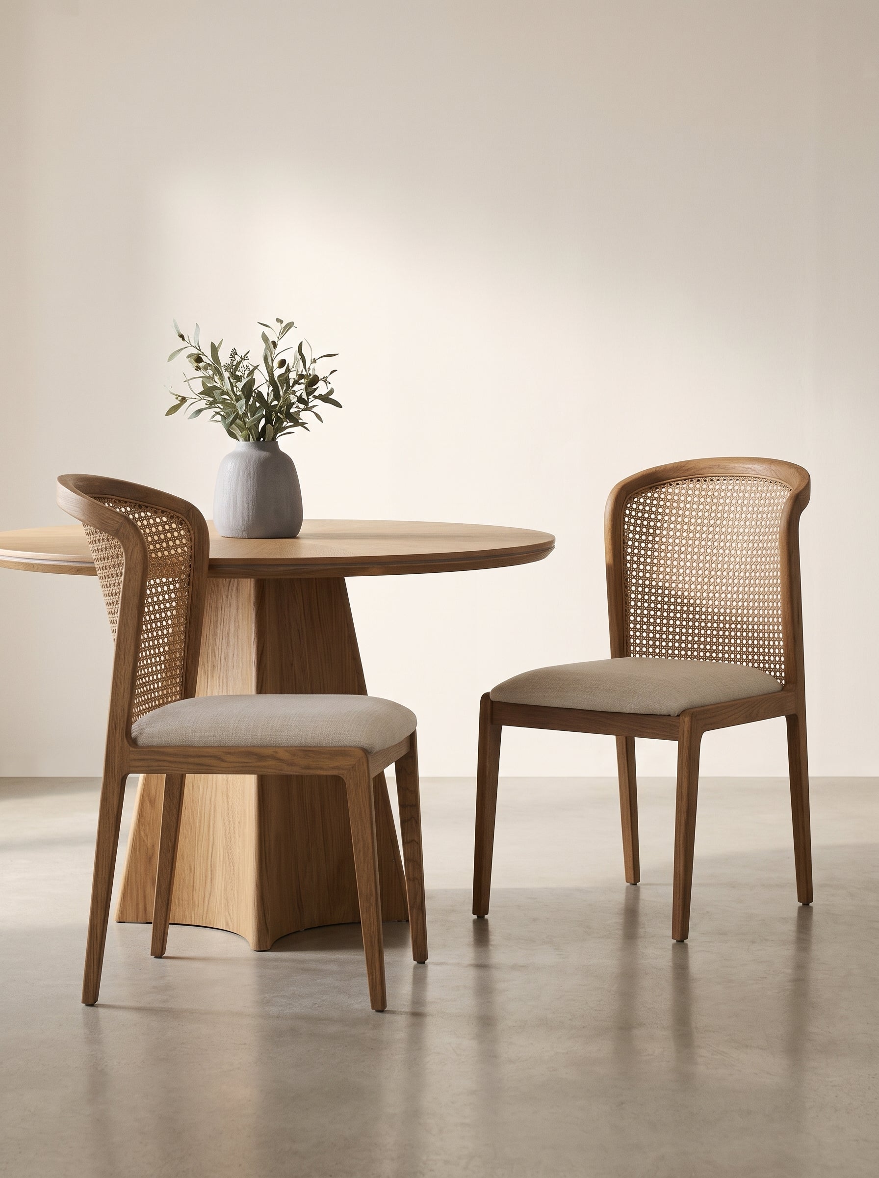 A minimalist dining set featuring a round light oak table and two matching wooden chairs with woven cane backs and light beige upholstered seats, styled with a gray ceramic vase of greenery on a neutral beige background.