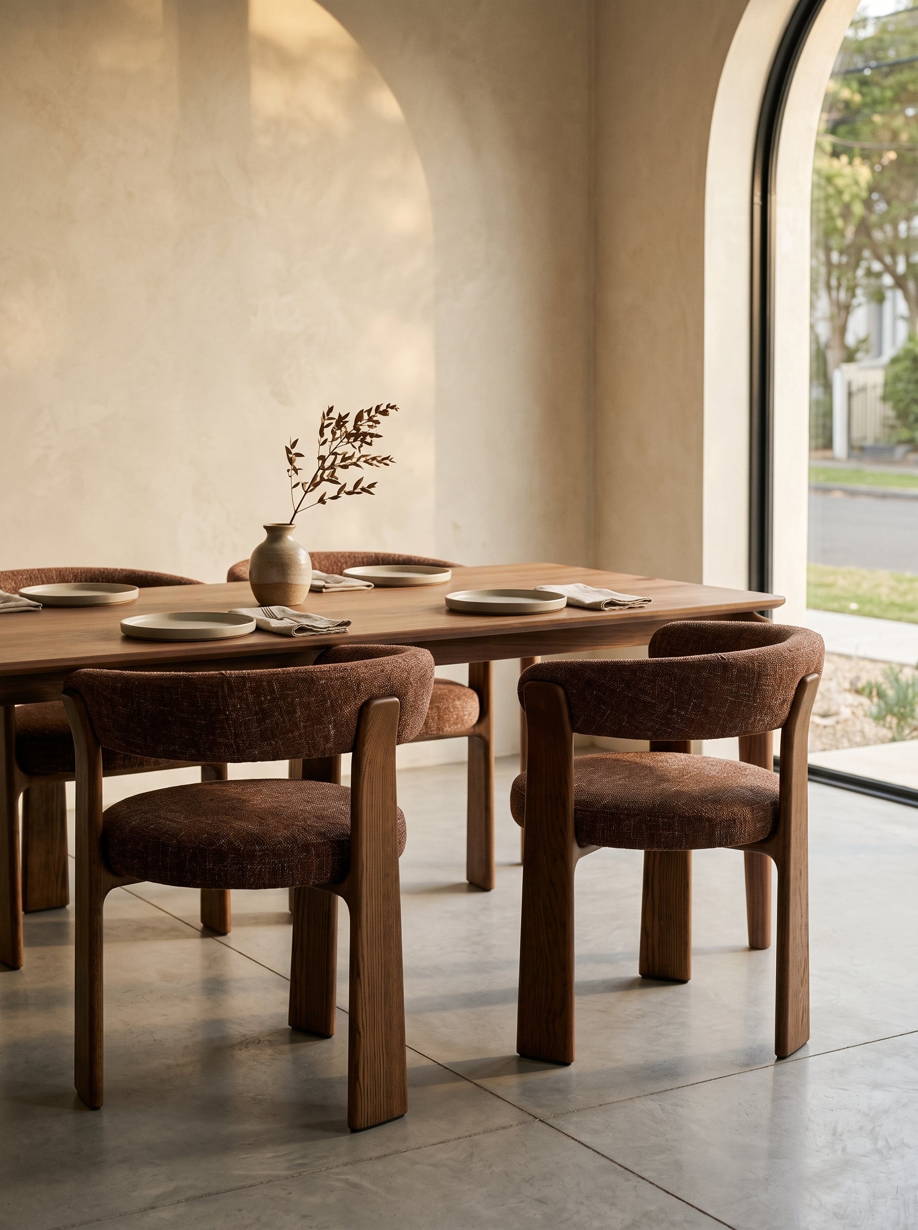 A modern dining room scene featuring a wooden dining table and four upholstered dining chairs in a warm brown tone, set against a textured beige wall with a large arched window.