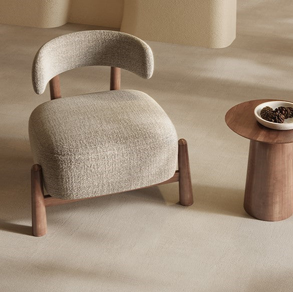A modern upholstered accent chair with a curved backrest and tapered wooden legs, shown beside a small round wooden side table in a warm neutral interior setting.