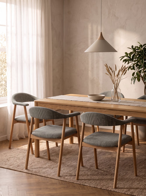 A dining room scene with a light wood rectangular table and six upholstered chairs in a soft gray fabric, arranged on a woven rug under a pendant light in a warm neutral interior.