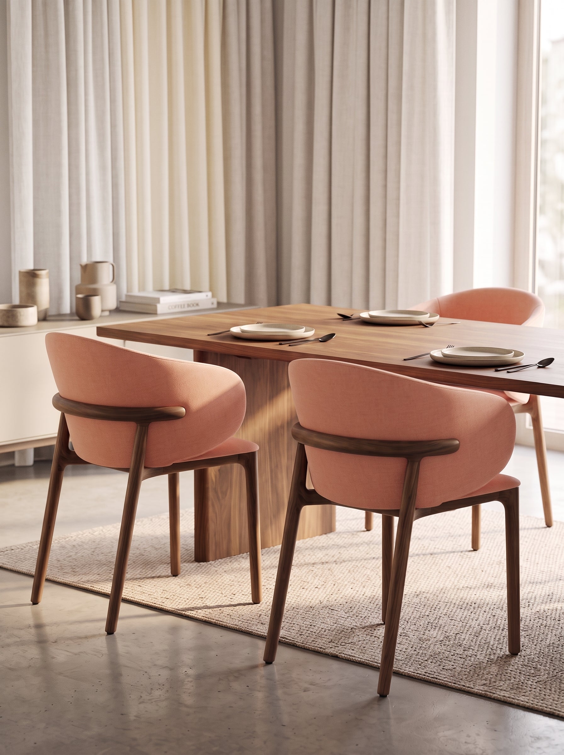A modern dining room scene featuring a rectangular wooden table and four upholstered dining chairs in a soft blush pink color with dark wood frames, set on a textured beige rug.