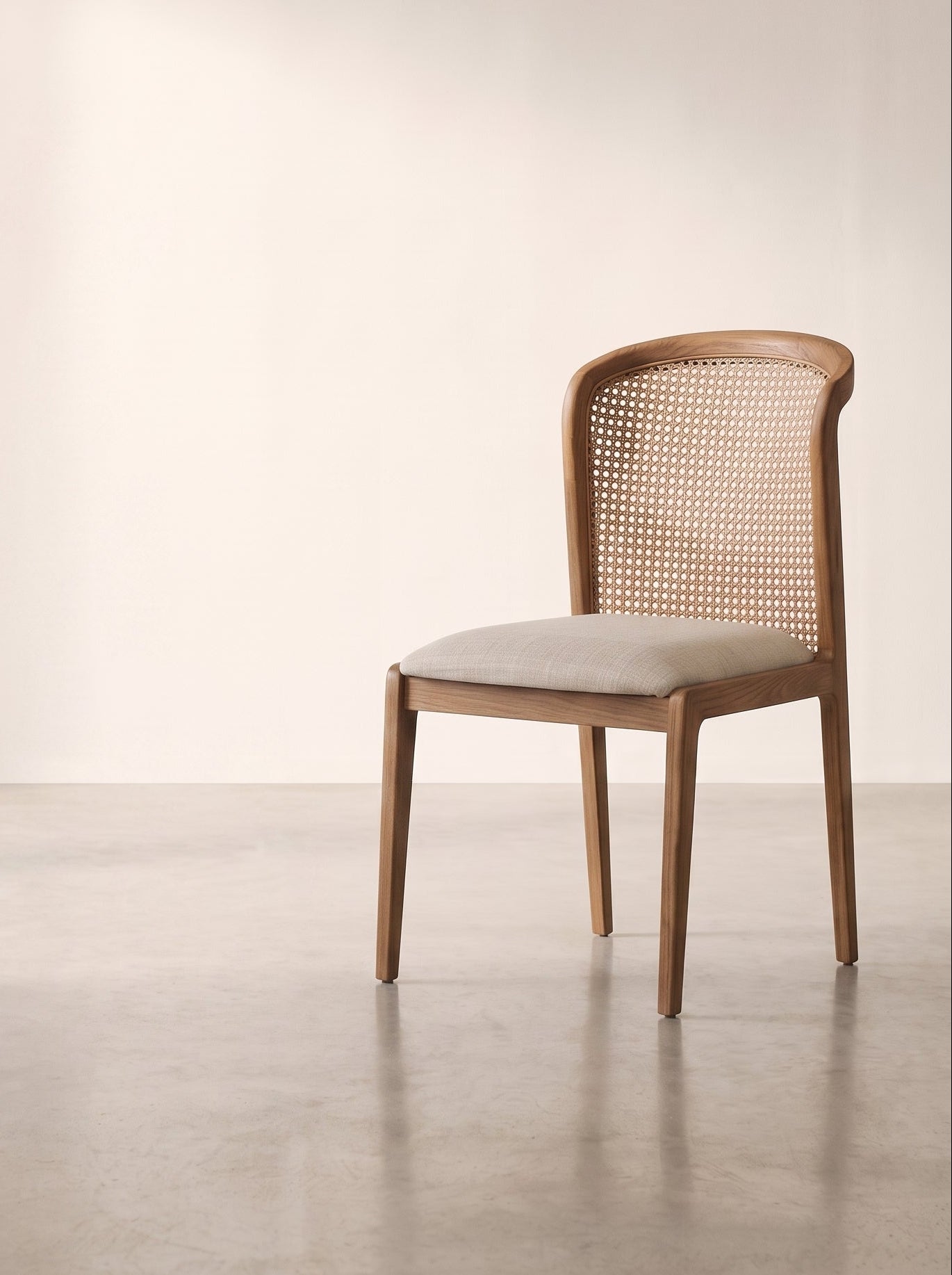 A modern wooden dining chair with a woven cane backrest and a light beige upholstered seat, positioned against a minimalist beige wall and concrete floor.